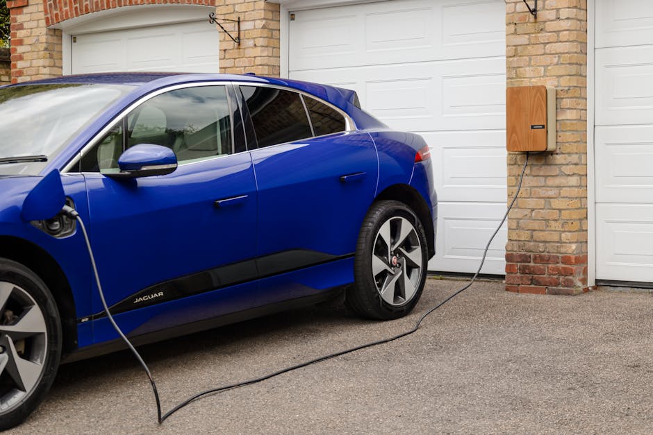 A blue Jaguar electric car is parked outside a residential property on a paved driveway, connected to a wall-mounted charging point with an black charging cable. The vehicle's side mirror and windows are visible, with the front left door closed and the rear left door slightly visible. The background features a brick wall and a white sectional garage door, indicating a typical home entrance for relocation purposes. The scene appears to be during daytime with natural lighting, and the area is tidy and unobstructed, suitable for home relocation or furniture transport activities as coordinated by Man and Van Rotherhithe.