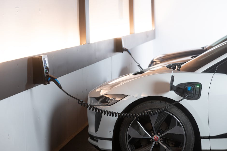 The image shows a white electric car parked in an indoor charging station, connected to a wall-mounted charging unit with a black charging cable. The car's front right side, including the headlight, wheel, and charging port, is visible, with the cable plugged securely into the port. The charging station is mounted on a gray wall, with a power socket and cable management visible. Natural light illuminates the area through windows above, and the scene reflects a clean, organized environment associated with home relocation or furniture transport involving electric vehicle charging during a moving process. This setting is representative of logistics and transportation aspects in house removals, notably when managing electric vehicles as part of a move, with the work area free of clutter and equipment, aligning with professional moving services such as those provided by Man and Van Rotherhithe.