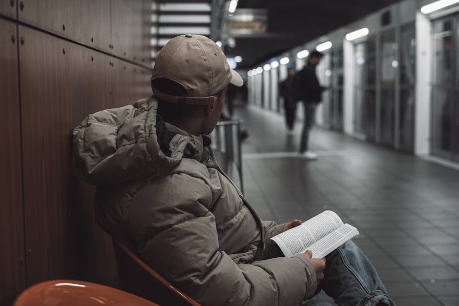 A person wearing a light grey cap and a puffy beige jacket is seated on an orange chair against a wooden-paneled wall inside a modern, well-lit train station. They are holding and reading a book with a white cover and black text. In the background, a blurred figure is walking along the platform, which is lined with glass panels and metallic structures, indicating an active setting for home relocation and moving services. The environment suggests a waiting area or transit point, with a focus on preparation for furniture transport or packing before a house move, as referenced in the page about book removals near Canada Water Station with local experts, Man and Van Rotherhithe.