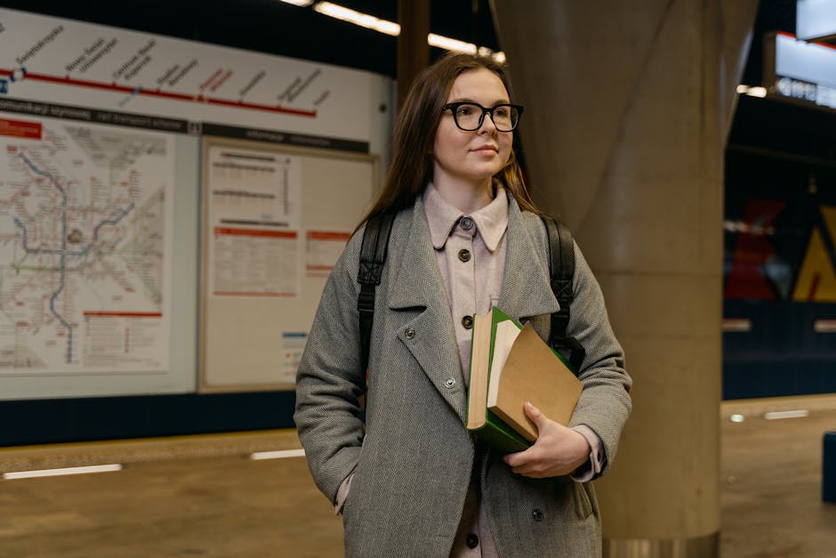A young woman with long brown hair, wearing glasses, a light grey coat, and carrying a black backpack, stands inside a metro station near a large map display. She holds several books and folders, including a green-covered notebook, in her left hand, pressed against her torso. The background features a detailed subway map with red and black lines, signage at the station, and a concrete column supporting the ceiling. The lighting is artificial and evenly distributed across the scene, illustrating a typical setting for a home relocation or moving process involving packing and transit logistics, as managed by Man and Van Rotherhithe. The image captures an environment where a client is preparing for a house move, highlighting the importance of organized packing and transportation, consistent with the services offered for removals near Canada Water Station.