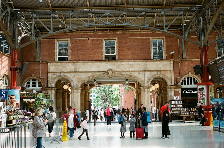A person wearing a light grey cap and a puffy beige jacket is seated on an orange chair against a wooden-paneled wall inside a modern, well-lit train station. They are holding and reading a book with a white cover and black text. In the background, a blurred figure is walking along the platform, which is lined with glass panels and metallic structures, indicating an active setting for home relocation and moving services. The environment suggests a waiting area or transit point, with a focus on preparation for furniture transport or packing before a house move, as referenced in the page about book removals near Canada Water Station with local experts, Man and Van Rotherhithe.
