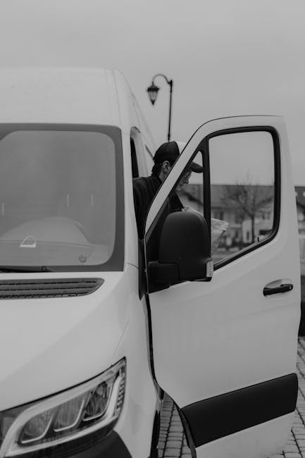 A man wearing a dark jacket and cap is partially visible inside a white cargo van with the driver's door open, positioned on a paved street in Rotherhithe. The van is parked near a lamppost and other buildings, with a row of leafless trees in the background, indicating a possible early morning or overcast day. Inside the van, visible packing materials include cardboard boxes and plastic wrapping, suggesting preparations for a house relocation. The open door reveals the interior space used for loading or unloading household items such as furniture, boxes, and packing supplies, which are arranged on the vehicle’s floor. The scene captures the loading process typical of professional removals, with the van’s exterior featuring a modern design and large side mirror. The setting reflects a typical urban environment suitable for furniture transport and moving services provided by companies like Man and Van Rotherhithe, as part of efficient home relocation logistics.