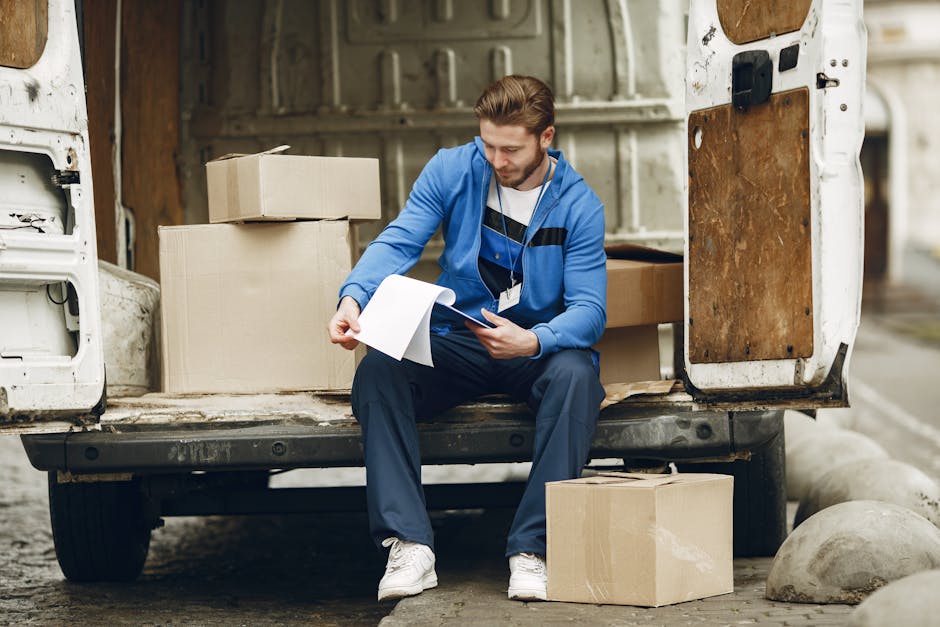 A man wearing a blue jacket and dark trousers is seated on the open tailgate of a white moving van, reviewing a clipboard and taking notes during a home relocation process. The van's interior is visible, with cardboard boxes of various sizes stacked inside, some wrapped in plastic or packing paper. The man is surrounded by additional cardboard boxes placed on the ground nearby, and the pavement beneath is partly covered with concrete bollards. The loading area appears to be outside a building during daytime, with natural light illuminating the scene. The image captures the logistics stage of furniture transport and packing as part of a professional removals service, such as those offered by Man and Van Rotherhithe, supporting efficient home moving and packing and moving processes.