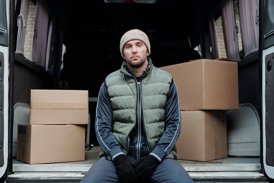 A man wearing a dark jacket and cap is partially visible inside a white cargo van with the driver's door open, positioned on a paved street in Rotherhithe. The van is parked near a lamppost and other buildings, with a row of leafless trees in the background, indicating a possible early morning or overcast day. Inside the van, visible packing materials include cardboard boxes and plastic wrapping, suggesting preparations for a house relocation. The open door reveals the interior space used for loading or unloading household items such as furniture, boxes, and packing supplies, which are arranged on the vehicle’s floor. The scene captures the loading process typical of professional removals, with the van’s exterior featuring a modern design and large side mirror. The setting reflects a typical urban environment suitable for furniture transport and moving services provided by companies like Man and Van Rotherhithe, as part of efficient home relocation logistics.