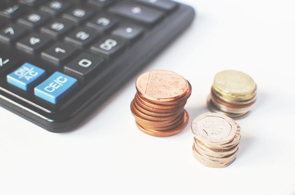 Close-up image showing a calculator with blue and gray buttons positioned in the top-left corner, and three separate stacks of British coins — one copper-colored, one silver-colored, and one gold-co