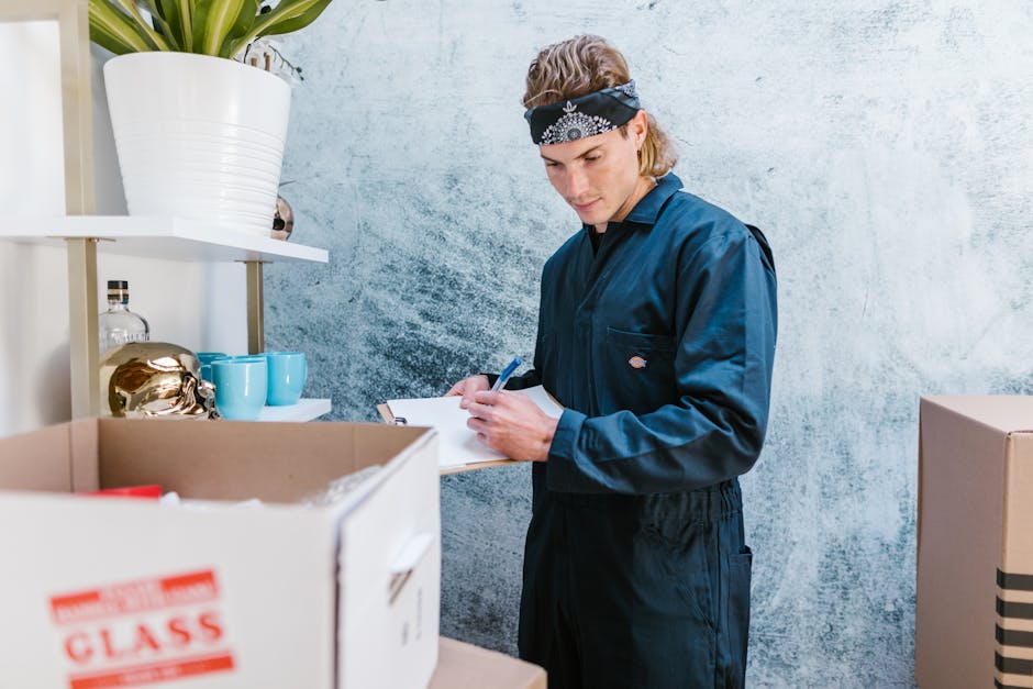 A young man wearing a dark blue work jumpsuit and a black bandana with white patterns, standing indoors beside a cluttered area of packing boxes and shelves, is seen writing on a clipboard with a pen. The scene is set against a textured light grey wall, with a white shelf holding a large white pot with a green plant, ceramic cups, and decorative items such as a gold-colored object and glass bottles. In the foreground, there are open cardboard boxes, one labeled 'CLASS', containing packing materials or household items, indicating an organized packing or home relocation process. The lighting is natural, illuminating the workspace where furniture and belongings are being prepared for transport, aligning with professional removals and moving services offered by Man and Van Rotherhithe as part of their furniture transport and packing and moving logistics, supporting their expertise in house removals near Greenland Dock.