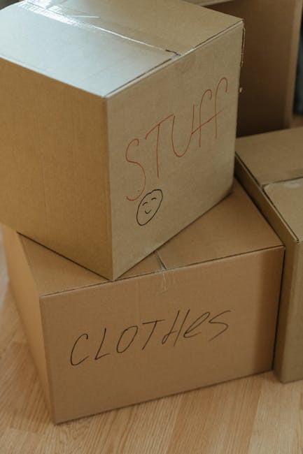 Close-up image of three plain cardboard boxes stacked indoors, used for house removals and packing during a home relocation. The top box has handwritten text in red marker that reads 'STUFF' with a small, smiling face drawn underneath. The box below it is labeled 'CLOTHES' also in red marker. The boxes are placed on a wooden floor near other similar boxes and packaging materials, indicating an organised packing process. The setting suggests preparation for furniture transport, packing and moving, or loading into a van, with natural lighting emphasizing the inventory organization by Man and Van Rotherhithe, a professional removals service provider.
