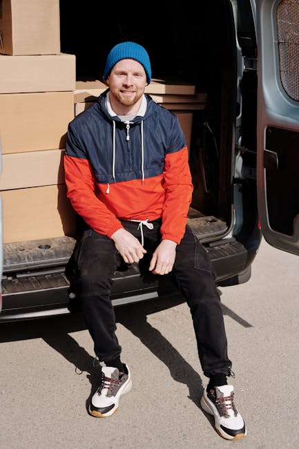 A man wearing a blue beanie, navy and red waterproof jacket, and black trousers is sitting on the open back door of a black moving van. Inside the van, there are several large cardboard boxes stacked neatly. The boxes vary in size and are secured with packing tape. The man is smiling and appears relaxed, with his hands resting on his knees. The van is parked outdoors on a paved surface, with part of a residential building visible in the background. The sliding side door of the van is open, revealing additional packing materials such as plastic wrap and blankets used for furniture protection during the home relocation process. This scene depicts the loading stage of a furniture transport and packing operation conducted by Man and Van Rotherhithe, assisting with house removals and moving services in the Rotherhithe area.