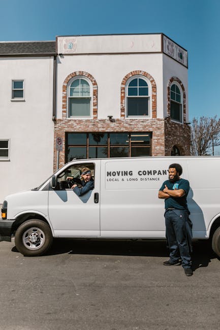A white moving van parked on the street in front of a two-storey building with white and brick exterior walls and arched windows on the upper floor. The van has branding that reads 'MOVING COMPANY LOCAL & LONG DISTANCE' and is positioned near the building entrance, suggesting preparation for house relocation. Inside the driver’s window, a person wearing a cap and glasses is seen talking to a colleague standing outside the van. The colleague, dressed in dark clothing with arms crossed, is standing on the pavement next to the vehicle, engaged in conversation. The scene is lit by natural daylight, with a clear blue sky overhead, indicating a typical moving day setup involving furniture transport, packing, and loading processes for home relocation services. The image highlights professional moving activity carried out by Man and Van Rotherhithe, focused on safe and efficient moving logistics.