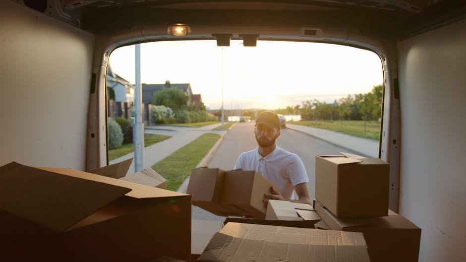 A man wearing a blue beanie, navy and red waterproof jacket, and black trousers is sitting on the open back door of a black moving van. Inside the van, there are several large cardboard boxes stacked neatly. The boxes vary in size and are secured with packing tape. The man is smiling and appears relaxed, with his hands resting on his knees. The van is parked outdoors on a paved surface, with part of a residential building visible in the background. The sliding side door of the van is open, revealing additional packing materials such as plastic wrap and blankets used for furniture protection during the home relocation process. This scene depicts the loading stage of a furniture transport and packing operation conducted by Man and Van Rotherhithe, assisting with house removals and moving services in the Rotherhithe area.