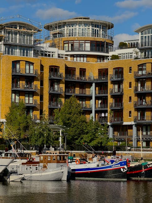 A quayside scene with several boats docked along the water, including a white boat, a blue and white boat named Leone, and a black boat, all secured to the quay. Behind the boats, there is an apartment complex with multiple floors featuring yellow brickwork, glass balconies, and large windows. The building has a distinctive modern design with curved structures on the upper levels and exterior scaffolding indicating ongoing construction or renovation. Green trees are visible at the base of the building, partially obscuring some lower floors. The scene is set during daytime with a partly cloudy sky, and the water reflects the boats and buildings. This image relates to home and furniture transport, similar to house removals services offered by Man and Van Rotherhithe, with the context of moving logistics and planning for flat relocations without lift access.
