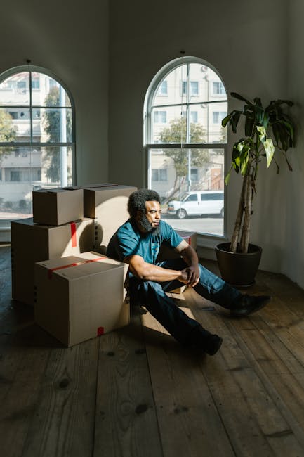 A man with curly dark hair, wearing a blue T-shirt and dark trousers, sits on a wooden floor inside a room with large, arched windows that let in natural light. He is surrounded by several cardboard boxes, some sealed with red tape and others open, revealing packing materials. The boxes are stacked and positioned around him, indicating a home relocation process. A tall potted plant is located near the window on the right side of the image. Outside the windows, a street scene with parked cars and residential buildings is visible. The room appears to be uncluttered, with a simple, clean environment, typical of a house being prepared for moving or packing. This scene illustrates the packing or loading stage in a furniture transport or house removal service, with clear focus on the boxes and the interior space, aligning with moving and relocation services provided by Man and Van Rotherhithe.