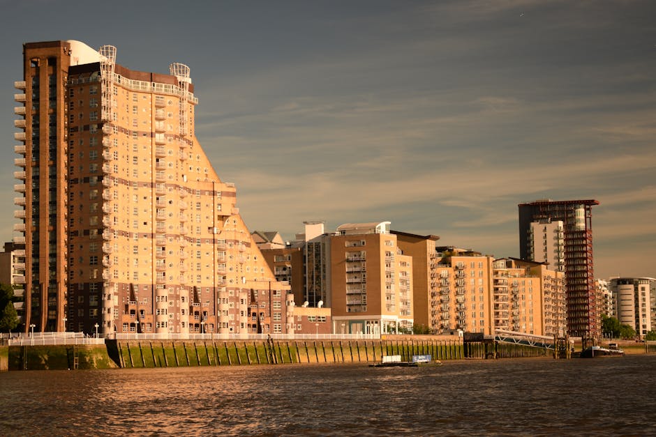A quayside scene with several boats docked along the water, including a white boat, a blue and white boat named Leone, and a black boat, all secured to the quay. Behind the boats, there is an apartment complex with multiple floors featuring yellow brickwork, glass balconies, and large windows. The building has a distinctive modern design with curved structures on the upper levels and exterior scaffolding indicating ongoing construction or renovation. Green trees are visible at the base of the building, partially obscuring some lower floors. The scene is set during daytime with a partly cloudy sky, and the water reflects the boats and buildings. This image relates to home and furniture transport, similar to house removals services offered by Man and Van Rotherhithe, with the context of moving logistics and planning for flat relocations without lift access.
