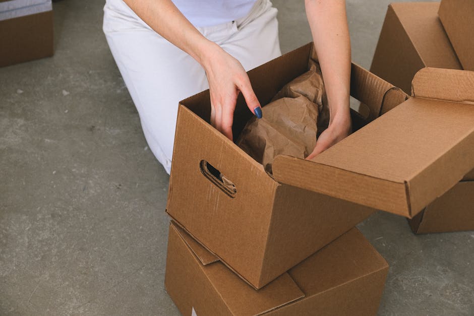 Close-up of a person dressed in white trousers and a light-colored top, packing small items into a brown cardboard moving box lined with crumpled brown packing paper, with additional closed boxes surrounding them on a concrete floor. The individual's hands are actively placing or removing the packing materials, highlighting a packing and moving process typical for home relocation. The scene is set indoors, outside a doorway or on a pavement area, with natural lighting illuminating the area. This image illustrates the preparation and careful packing involved in furniture transport and residential removals, a service offered by Man and Van Rotherhithe. Visible elements include cardboard boxes, packing paper, and the person's hands working to secure belongings for a transfer to a moving van or storage area.