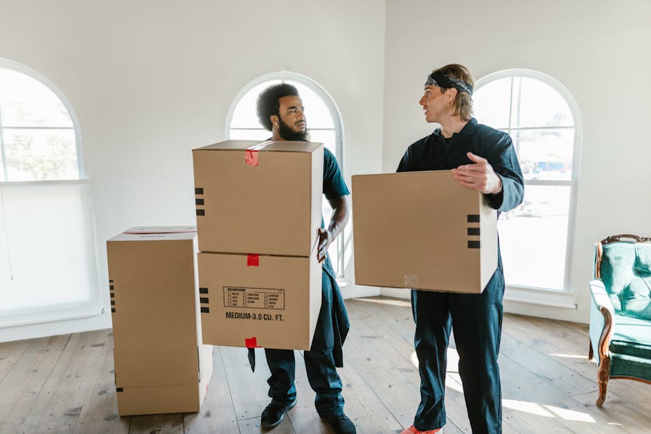 Two professional movers from Man and Van Rotherhithe are inside a bright room with natural light coming through arched windows. One mover, with a beard and curly hair, is holding two stacked cardboard boxes labeled for home relocation, while the other, wearing a headband, carries a single cardboard box. The boxes are sealed with red tape and have printed labels indicating size and content. There are additional boxes on the wooden floor nearby, some partially visible, suggesting packing for a furniture transport or moving process. The movers are dressed in dark clothing suitable for loading and packing tasks, and the room features light-colored walls with minimal furniture, including a green armchair with wooden accents positioned on the right side. This scene illustrates the careful handling and loading of packing materials during the loading process of a removal service, reflecting efficient packing and transport logistics. Man and Van Rotherhithe provides professional removals supporting house moves with proper packing, loading, and transportation.