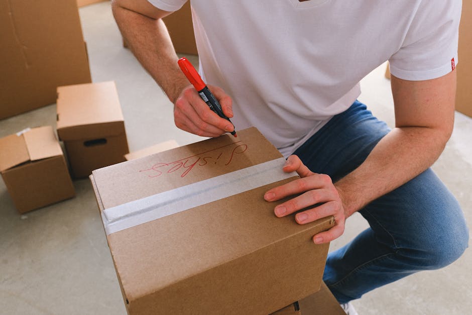 Close-up of a person dressed in white trousers and a light-colored top, packing small items into a brown cardboard moving box lined with crumpled brown packing paper, with additional closed boxes surrounding them on a concrete floor. The individual's hands are actively placing or removing the packing materials, highlighting a packing and moving process typical for home relocation. The scene is set indoors, outside a doorway or on a pavement area, with natural lighting illuminating the area. This image illustrates the preparation and careful packing involved in furniture transport and residential removals, a service offered by Man and Van Rotherhithe. Visible elements include cardboard boxes, packing paper, and the person's hands working to secure belongings for a transfer to a moving van or storage area.