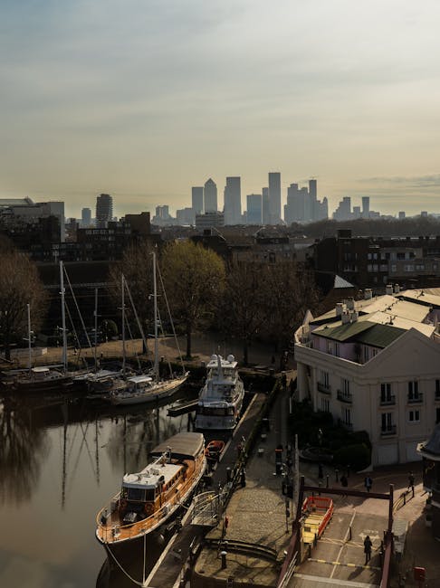 An aerial view showing a marina with several moored boats and yachts, some covered with protective tarps, along a canal or river in Rotherhithe. In the foreground, a sidewalk runs parallel to the water, with a few pedestrians walking and a small red and yellow vehicle parked nearby. To the right, residential buildings with white facades and large windows are visible, indicating a residential area suitable for ongoing home relocations. In the background, the skyline of central London features tall office buildings and skyscrapers under a partly cloudy sky, suggesting a typical cityscape scene. The environment appears calm, with natural daylight illuminating the scene, suitable for logistical planning involved in furniture transport and packing during house removals. This image reflects the organized loading process of belongings onto transport vehicles by [COMPANY_NAME], such as Man and Van Rotherhithe, supporting services like removals and home relocation in the Rotherhithe area.