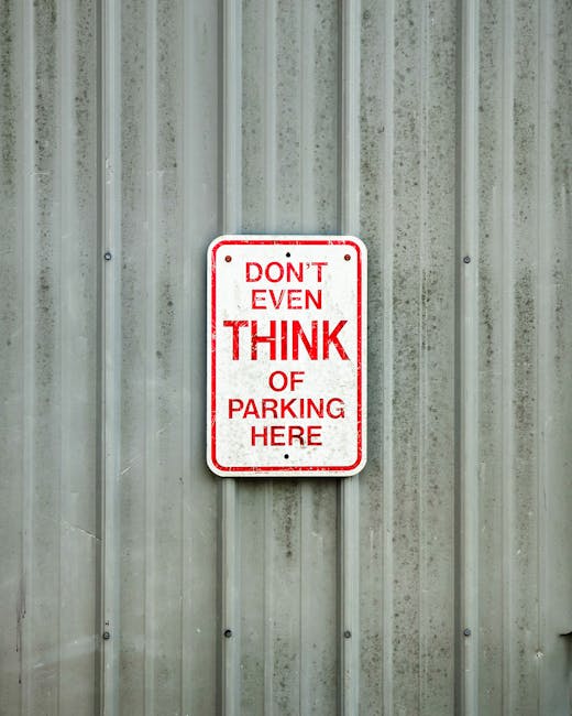 A rectangular metal sign with a white background and red text is mounted on a vertically ridged, metallic wall. The sign reads 'DON'T EVEN THINK OF PARKING HERE' in bold uppercase letters. The wall surface has visible bolts and a slightly weathered appearance. This image is related to house removals and moving services in Rotherhithe, as it may depict parking restrictions relevant for logistical planning during home relocation or furniture transport. The photo emphasizes the importance of adhering to parking rules under Southwark Council regulations for efficient loadings needed by Man and Van Rotherhithe’s removals service, illustrating the type of signage encountered during the loading process at properties situated within the area.