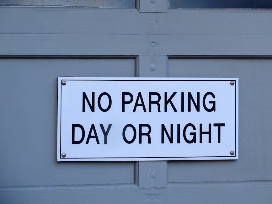 A rectangular metal sign with a white background and red text is mounted on a vertically ridged, metallic wall. The sign reads 'DON'T EVEN THINK OF PARKING HERE' in bold uppercase letters. The wall surface has visible bolts and a slightly weathered appearance. This image is related to house removals and moving services in Rotherhithe, as it may depict parking restrictions relevant for logistical planning during home relocation or furniture transport. The photo emphasizes the importance of adhering to parking rules under Southwark Council regulations for efficient loadings needed by Man and Van Rotherhithe’s removals service, illustrating the type of signage encountered during the loading process at properties situated within the area.
