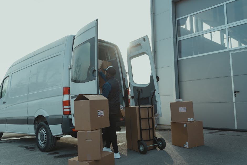 A worker at Man and Van Rotherhithe is seen during a home relocation process, standing beside an open white delivery van with its rear doors fully open, revealing packed cardboard boxes and wrapped furniture inside. Several additional cardboard boxes, some sealed with packing tape and labeled, are placed on the pavement nearby, ready for loading or unloading. The worker is wearing casual clothing, including a cap and gloves, and appears to be lifting or adjusting an item inside the vehicle. A hand truck or trolley is positioned on the pavement next to the van, used for transporting heavier boxes. The scene takes place outside a modern building with large glass windows and aluminum panels, with bright natural lighting suggesting daytime. This image captures the logistics involved in furniture transport and packing during a house removal, illustrating the careful handling, organized packing, and loading process supported by professional removals services like Man and Van Rotherhithe.