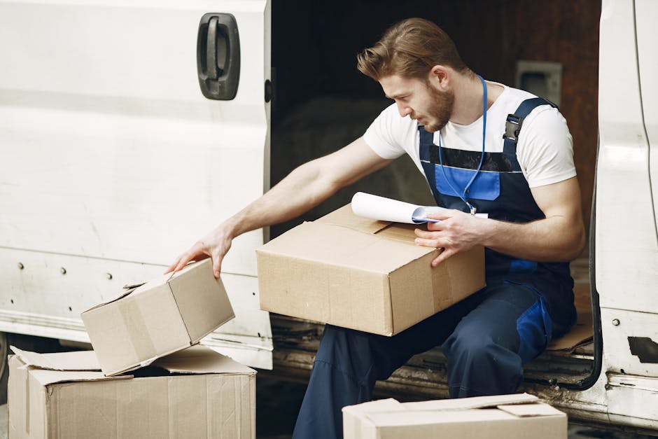A young man with long hair, wearing a dark blue work jumpsuit, casual sneakers, and a patterned headband, is sitting on a wooden floor inside a room with large, arched windows. He is surrounded by several cardboard boxes, some sealed with red packing tape, indicating packing for a home relocation or moving process. The room is well-lit by natural sunlight streaming through the windows, which reveal a view of neighboring buildings and trees outside. The man appears relaxed, possibly taking a break during the loading or packing phase of furniture transport and moving services with Man and Van Rotherhithe. The scene captures the preparation stage of a house removal, emphasizing packaging and handling of belongings, characteristic of professional removals tasks.