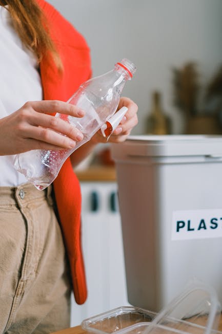 A person wearing a red cardigan and beige trousers holds a clear plastic bottle with a red cap, preparing to dispose of it into a recycling bin labeled 'PLASTIC' in a domestic setting. The vegetable-based waste management container is located indoors, positioned on a wooden surface, with a plastic packaging tray and other recyclable materials nearby. In the background, there is a washing machine and a neutral-colored wall, indicating a kitchen or utility area. The focus is on the individual’s hand and the plastic bottle, emphasizing proper recycling practices as part of home relocation or moving logistics managed by Man and Van Rotherhithe, which supports responsible waste disposal during removals.