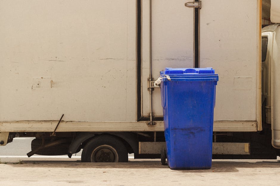 A large white panel van with a side door partially open, revealing a glimpse of the interior. Outside the van, a blue wheelie bin made of plastic is positioned on the pavement, secured with a white strap around its lid. The bin is located next to the rear wheel of the van, and its presence suggests waste disposal related to packing or remnants from a home relocation process. The setting appears to be outdoors, on a street or driveway, with natural lighting illuminating the scene. The van's exterior shows some signs of wear and weathering, and the pavement beneath is concrete. This image illustrates the stages of packing and waste removal involved in furniture transport and house removals, as managed by [COMPANY_NAME], supporting proper disposal rules for removals in Rotherhithe and SE16.