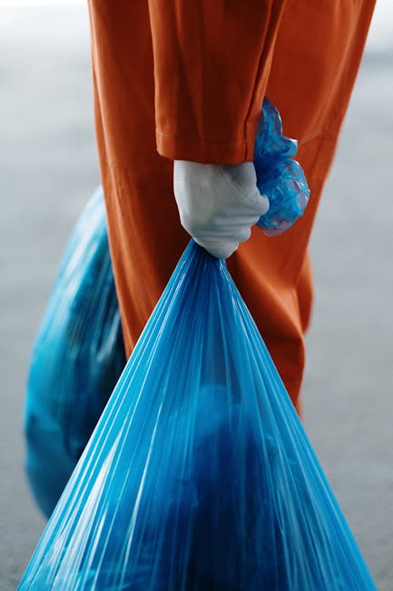 A person wearing a red cardigan and beige trousers holds a clear plastic bottle with a red cap, preparing to dispose of it into a recycling bin labeled 'PLASTIC' in a domestic setting. The vegetable-based waste management container is located indoors, positioned on a wooden surface, with a plastic packaging tray and other recyclable materials nearby. In the background, there is a washing machine and a neutral-colored wall, indicating a kitchen or utility area. The focus is on the individual’s hand and the plastic bottle, emphasizing proper recycling practices as part of home relocation or moving logistics managed by Man and Van Rotherhithe, which supports responsible waste disposal during removals.